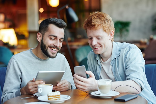 Twee gelukkige jongens met mobiele gadgets kijken naar nieuwsgierige dingen in de smartphone van een van hen terwijl ze tijd doorbrengen in café