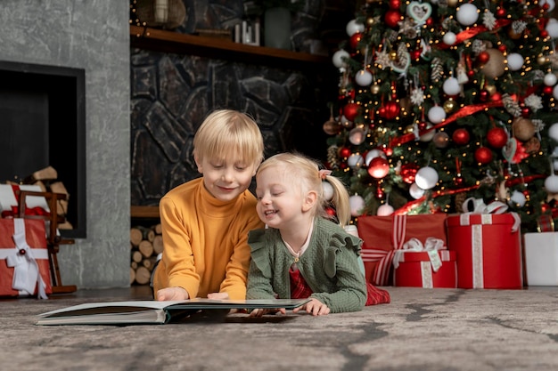 Twee gelukkige kinderen op de vloer onder de kerstboom. Blonde broer en zus lezen het boek kerstavond