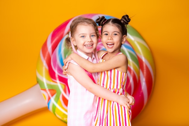 Twee gelukkige kleine meisjes in kleurrijke jurk lachen knuffelen plezier op gele ondergrond met lolly.