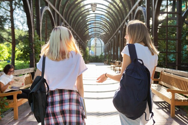 Twee gelukkige lachende pratende meisjes tieners studenten lopen samen, jonge vrouwen met rugzakken, zonnige dag op de achtergrond van het park, achteraanzicht