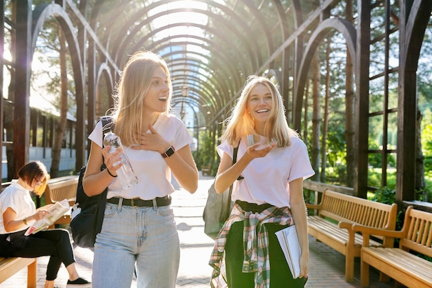 Twee gelukkige lachende pratende meisjes tieners studenten lopen samen, jonge vrouwen met rugzakken, zonnige dag op de achtergrond van het park