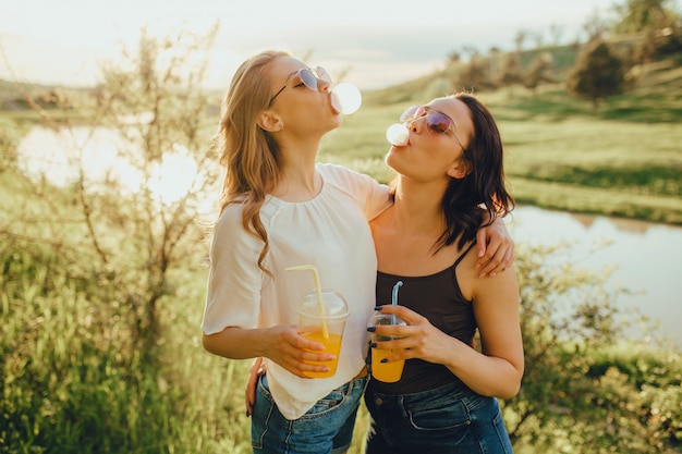 Twee gelukkige meisjes hebben plezier, blazen een bubbel op van een kauwgom, drinken cocktail uit de plastic beker met het rietje in de zonnebril, bij zonsondergang, positieve gezichtsuitdrukking, buiten