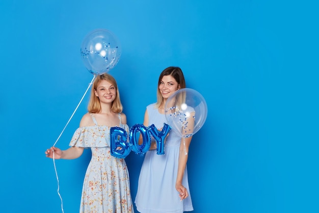 Twee gelukkige meisjes in jurk met blauwe ballonnen en ballon met de inscriptie jongen geïsoleerde blauwe achtergrond
