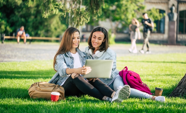 Twee gelukkige mooie jonge student vriendinnen in casual denim kleding zijn ontspannen in college park met laptop en smartphone door de universiteit en koffie drinken.
