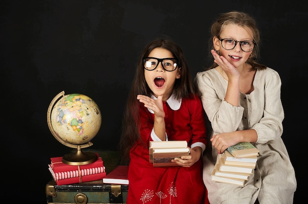 twee gelukkige mooie schoolmeisjes met globe en boeken zittend op de borst op een zwarte achtergrondgeluid