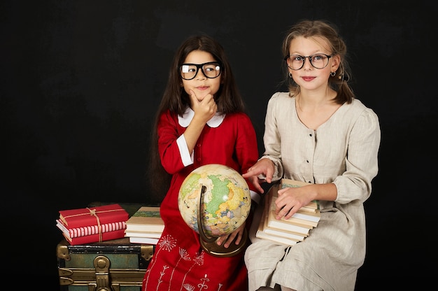 twee gelukkige mooie schoolmeisjes met globe en boeken zittend op de borst op een zwarte achtergrondgeluid