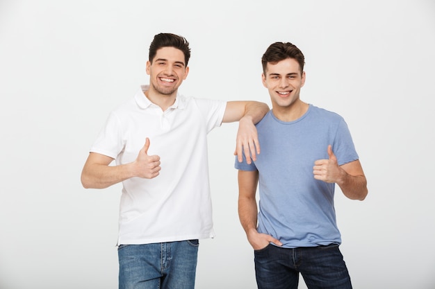 Twee gelukkige vrienden jaren '30 dragen van casual t-shirt en spijkerbroek glimlachend en duimen opdagen op camera in studio, geïsoleerd op witte achtergrond