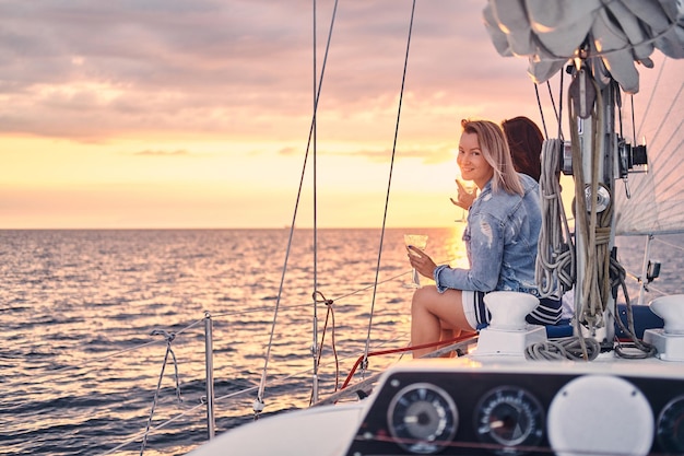 Twee gelukkige vriendinnen ontspannen op het jacht met glazen wijn in de handen, tijdens zonsondergang op volle zee.