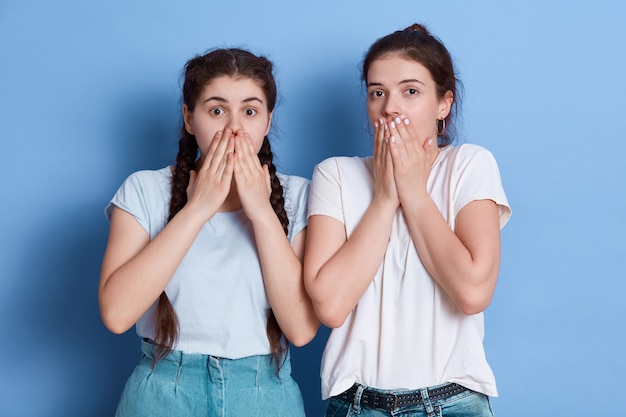 Twee geschokte vrouwen kleden zich met casual t-shirts die tegen een blauwe muur poseren die hun mond bedekt