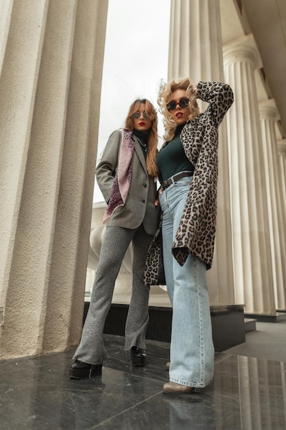 Twee glamoureuze jonge stijlvolle meisjes in modieuze kleding met modieuze schoenen staan en poseren in de buurt van de retro-kolom van het gebouw