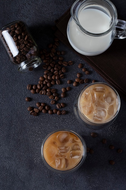 Twee glazen koude koffie met ijsblokjes, koffiebonen en kruik melk op een donkere achtergrond, bovenaanzicht