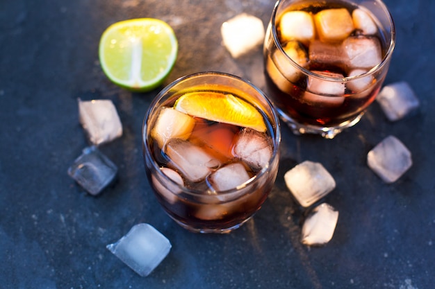 Twee glazen met cocktails: rum cola en cola whisky staan op een grijsblauwe betonnen gestructureerde achtergrond en ijsblokjes en gehakte limoen liggen rond