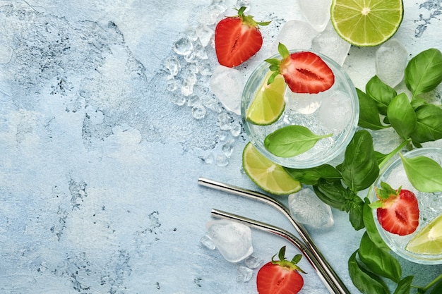 Twee glazen punch en verse ingrediënten voor het maken van limonade, infused detox water of cocktail. Aardbeien, limoen, munt, basilicum, ijsblokjes en shaker op grijze steen of betonnen ondergrond. Bovenaanzicht.