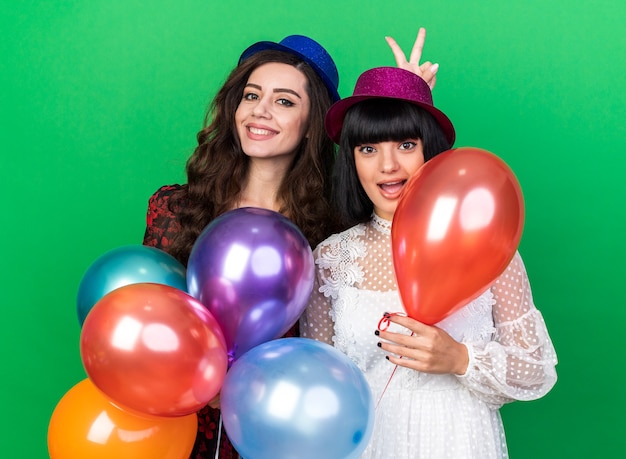 Twee glimlachende en onder de indruk jonge feestmeisjes met een feestmuts die beide ballonnen vasthoudt, een die konijnenoren maakt achter het hoofd van haar vriend geïsoleerd op een groene muur