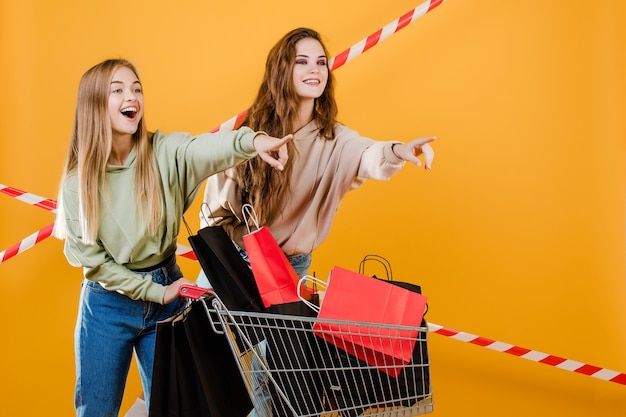 Twee glimlachende gelukkige mooie vrouwen die met karretje vingers richten op copyspace met kleurrijke die het winkelen zakken en signaalband over geel wordt geïsoleerd