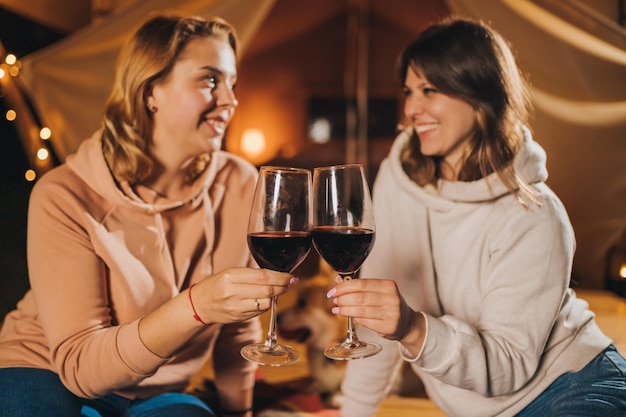 Twee glimlachende vrouwelijke vrienden die wijn drinken en fruit eten die in een gezellige glamping-tent zitten in de herfstavond vreugdevuur Luxe kampeertent voor buitenvakantie en vakantie Lifestyle-concept