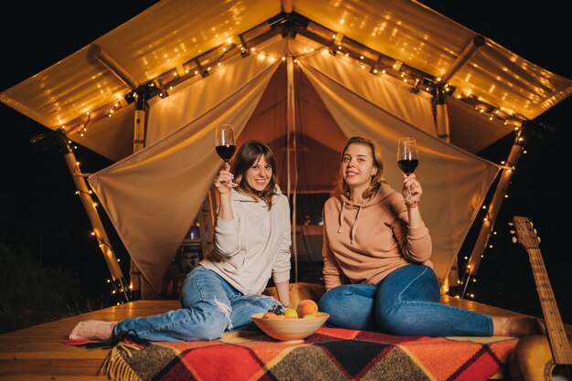 Twee glimlachende vrouwelijke vrienden die wijn drinken en fruit eten die in een gezellige glamping-tent zitten in de herfstavond vreugdevuur Luxe kampeertent voor buitenvakantie en vakantie Lifestyle-concept