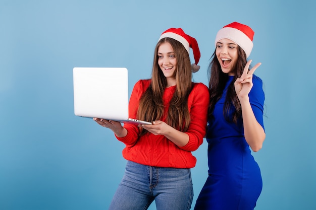 Twee glimlachende vrouwen die santahoeden dragen die laptop met gelukkige die gebaren bekijken over blauw worden geïsoleerd