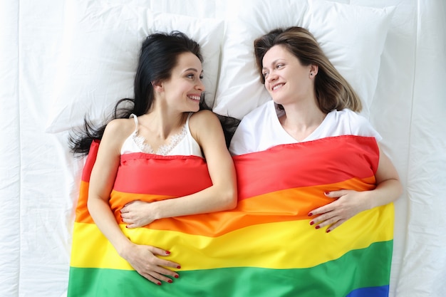 Twee glimlachende vrouwen liggen op bed met lgbt-vlag