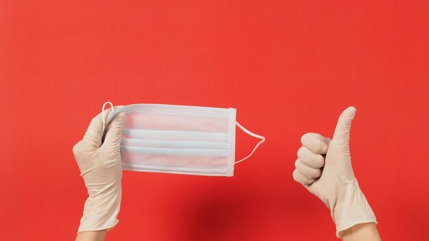 Twee handen dragen latexhandschoenen of witte medische handschoenen en gezichtsmasker op rode achtergrond.