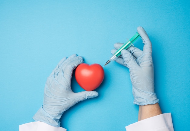 Twee handen in blauwe latex handschoenen houden rood hart en vastzittende naald van een plastic spuit erin op een blauwe achtergrond, bovenaanzicht