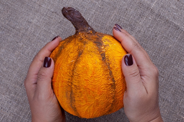 twee handen met een bruine manicure houden een zelfgemaakte oranje pompoen van papier-maché voor Halloween op een jute-oppervlak
