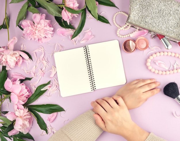 Twee handen met een zachte huid van een jong meisje en een handtas met cosmetica, boeket van bloeiende roze pioenrozen