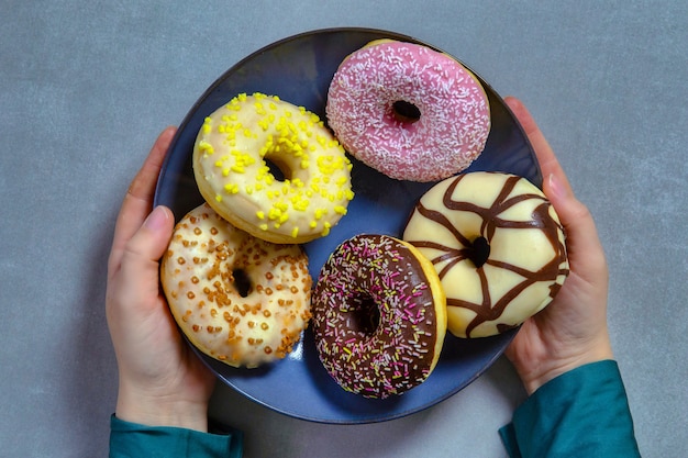 Twee handen met plaat van kleurrijke suikerachtige ronde geglazuurde donuts met gekleurde hagelslag op grijs