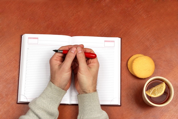 Twee handen met rode pen liggend op een notitieboekje op bureau met thee en koekjes biscuit