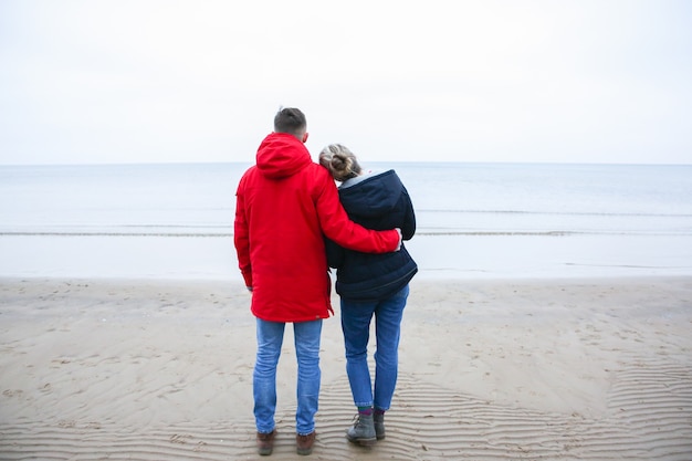 Twee hipsters die zich op het koude strand bevinden. Paar knuffelen en hand in hand. Liefdesverhaal in de buurt van de oceaan. Winterseizoen aan zee. Stijlvolle laarzen op het zand. Man in rode jas.