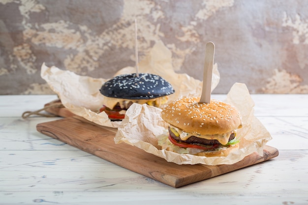 Twee huisgemaakte rundvleesburgers met donker en licht broodje, met greens, rode ui, tomaten, ingelegde komkommers en kaas in perkament op houten snijplank