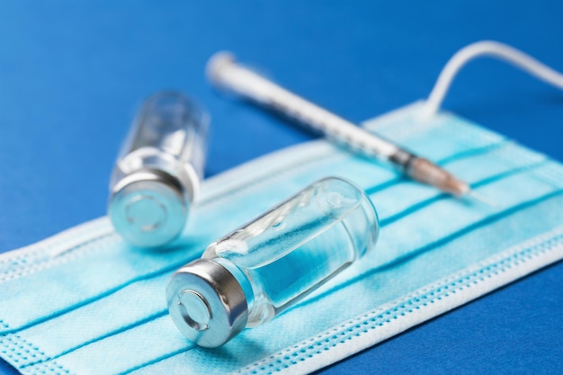 Twee injectieflacons met een oplossing voor injectie, een wegwerpspuit en een medisch masker op een blauwe achtergrond, vaccinatieconcept