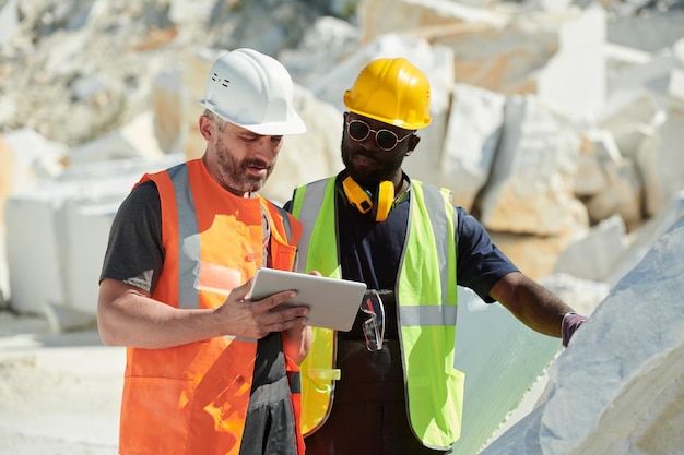 Twee interraciale mannelijke ingenieurs in veiligheidshelmen en uniform kijken naar tabletscherm