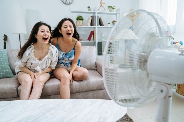 twee jonge Aziatische vriendinnen zittend op de bank van de bank in een gezellige woonkamer spelen met elektrische ventilator. speelse vrouwen die plezier hebben terwijl ze schreeuwen tegen de luchtkoeler in het appartement bij het raam in de zomer