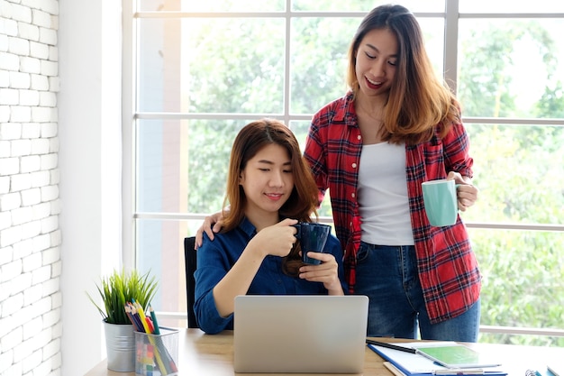 Twee jonge Aziatische vrouwen die koffiekop houden terwijl het werken met laptop computer