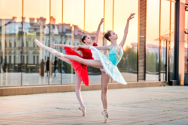 Twee jonge ballerina's in een felrode en blauwe tutu dansen tegen de achtergrond van de weerspiegeling van de stadszonsondergang