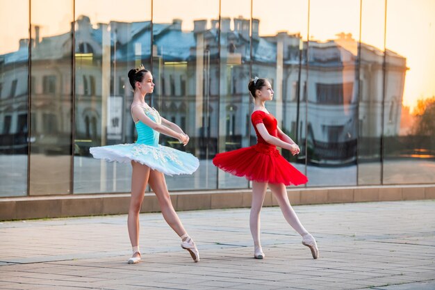 Twee jonge ballerina's in een felrode en blauwe tutu dansen tegen de achtergrond van de weerspiegeling van de stadszonsondergang