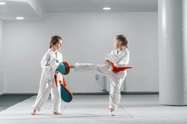 Twee jonge blanke meisjes in doboks met taekwondo training bij sportschool. Een meisje schopt terwijl een ander een schopdoel vasthoudt.