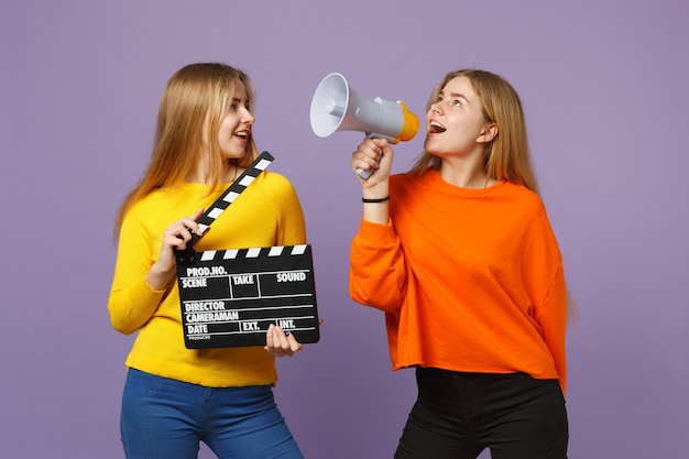 Twee jonge blonde tweelingzusters meisjes houden klassieke zwarte film filmklapper, schreeuwen op megafoon geïsoleerd op pastel violet blauwe muur. Mensen familie levensstijl concept.