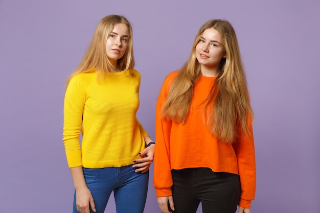 Twee jonge blonde tweelingzusters meisjes in levendige kleurrijke kleding staan, geïsoleerd op pastel violet blauwe muur. Mensen familie levensstijl concept.