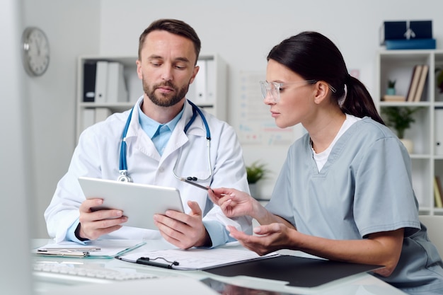 Twee jonge collega's in medisch uniform zitten aan een bureau in een medisch kantoor, scrollen door online informatie op tabletdisplay en raadplegen