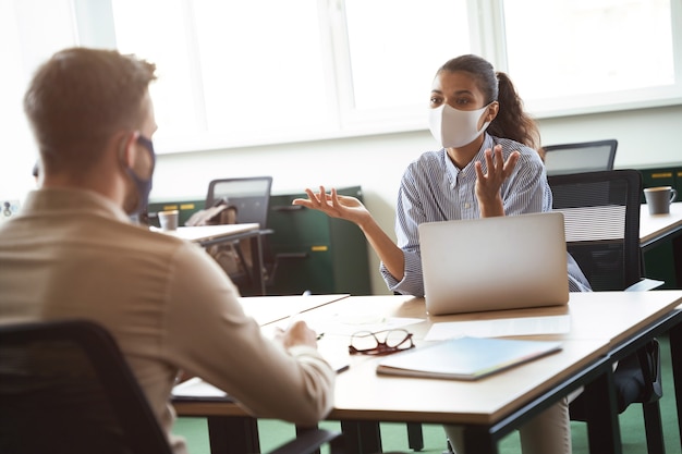 Twee jonge diverse collega's die een beschermend gezichtsmasker dragen die iets bespreken tijdens het werken