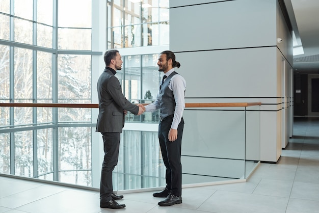 Twee jonge elegante zakenpartners die elkaar de hand schudden terwijl ze een deal sluiten in een groot kantoorgebouw