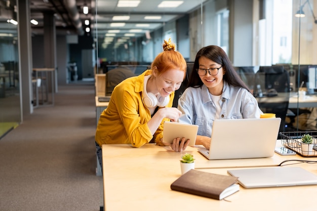 Twee jonge gelukkige interculturele zakenvrouwen kijken naar het tabletscherm dat door een van hen wordt vastgehouden en bespreken online grappige video op kantoor