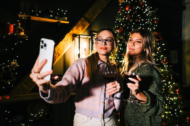 Foto twee jonge gelukkige vrouwen die een feestelijke foto-selfie maken in de buurt van de kerstboom en wijn drinken.