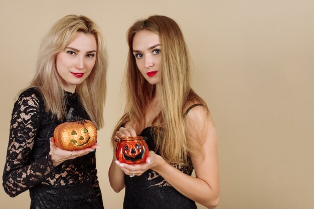 Twee jonge glimlachende vrouwen die Halloween-pompoenen op een beige achtergrond houden