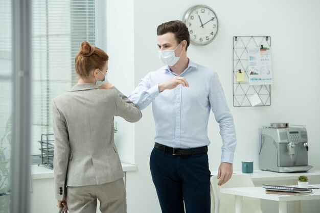 Twee jonge hedendaagse kantoormanagers in formele kleding en beschermende maskers die elkaar een elleboogstoot geven in plaats van handdruk of knuffel