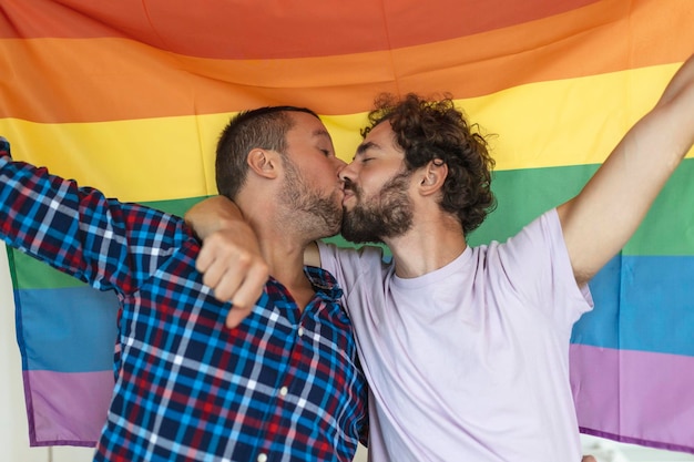 Foto twee jonge homoseksuele geliefden die elkaar liefdevol kussen twee jonge mannelijke geliefden die samen staan tegen een trotsvlag aanhankelijk jong homopaar die een romantisch moment samen delen