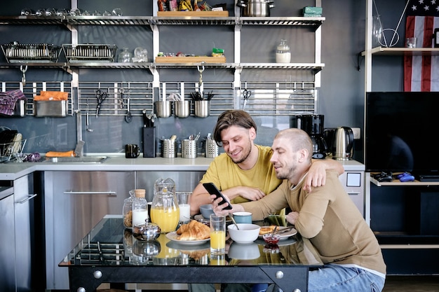 Twee jonge homoseksuele mannen knuffelen die op sociale media op smartphone scrollen tijdens het ontbijt aan de keukentafel