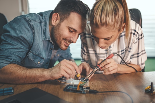 Foto twee jonge ingenieurs zijn gericht op het repareren van elektronische apparatuur door soldeerbout. man helpt vrouw solderen elementen van printplaat.